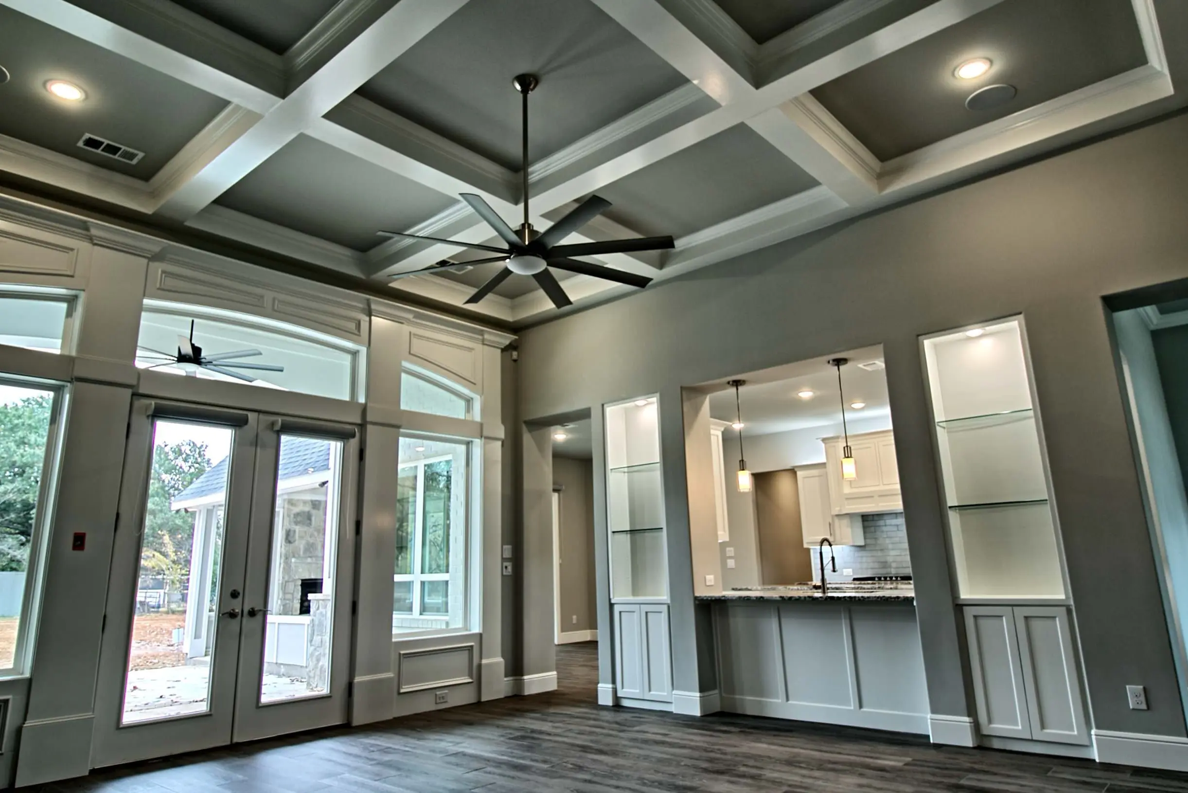 Living room with coffered ceiling, eight-blade ceiling fan, French doors and arched-transom windows to a covered patio, and a kitchen pass-through between lit glass-shelf niches, in North Richland Hills, TX