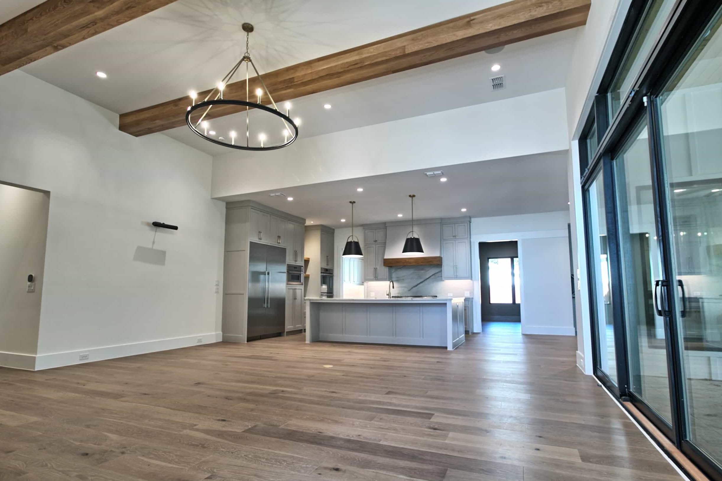 Great room with wood ceiling beams, black ring chandelier and wide-plank floors, open to a gray shaker kitchen with island, black pendants and black-framed sliding doors, in Colleyville, TX