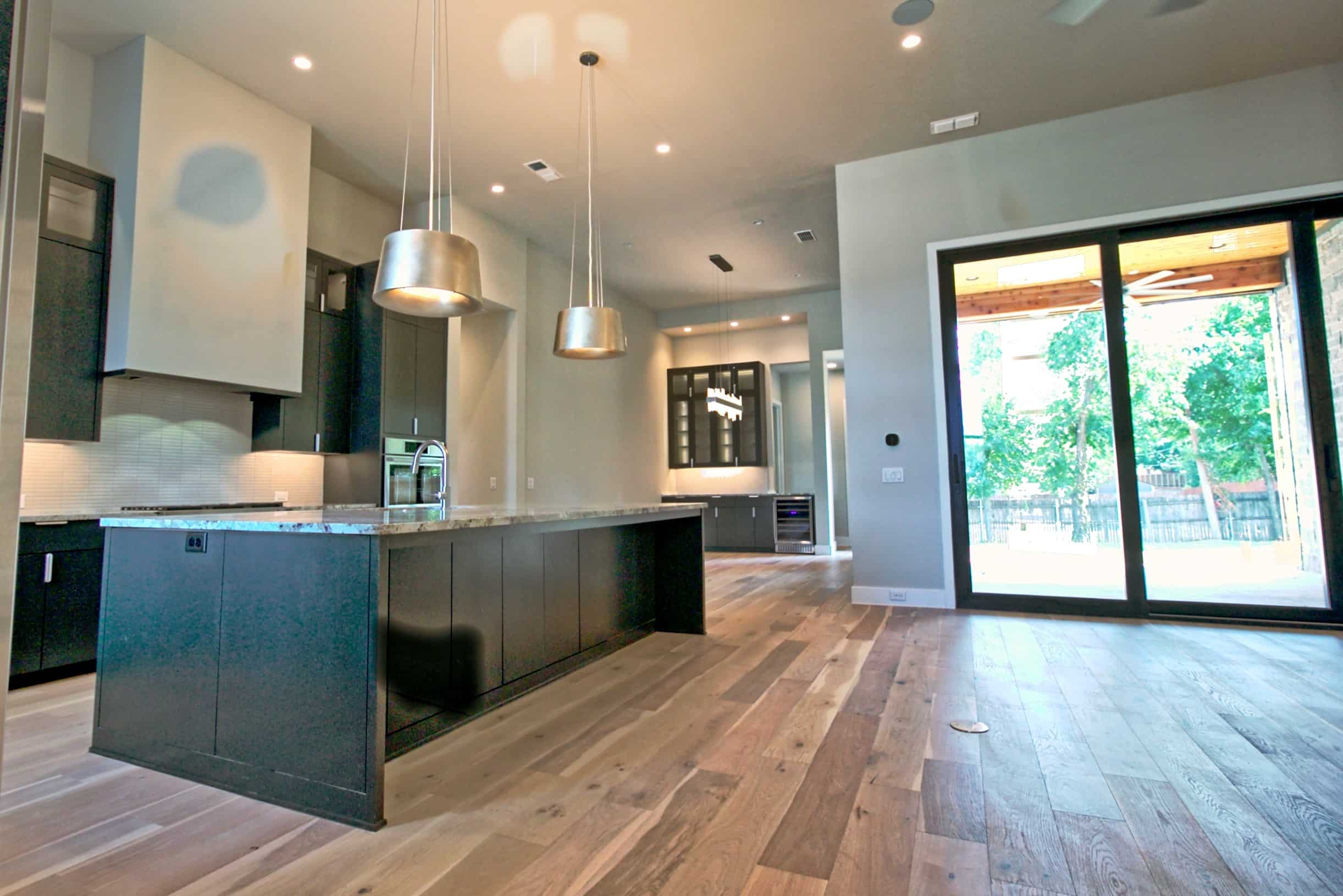 Open kitchen with charcoal flat-panel cabinetry, long granite-topped island, silver drum pendants, wide-plank wood floors and black-framed sliding doors to a covered patio, in North Richland Hills, TX