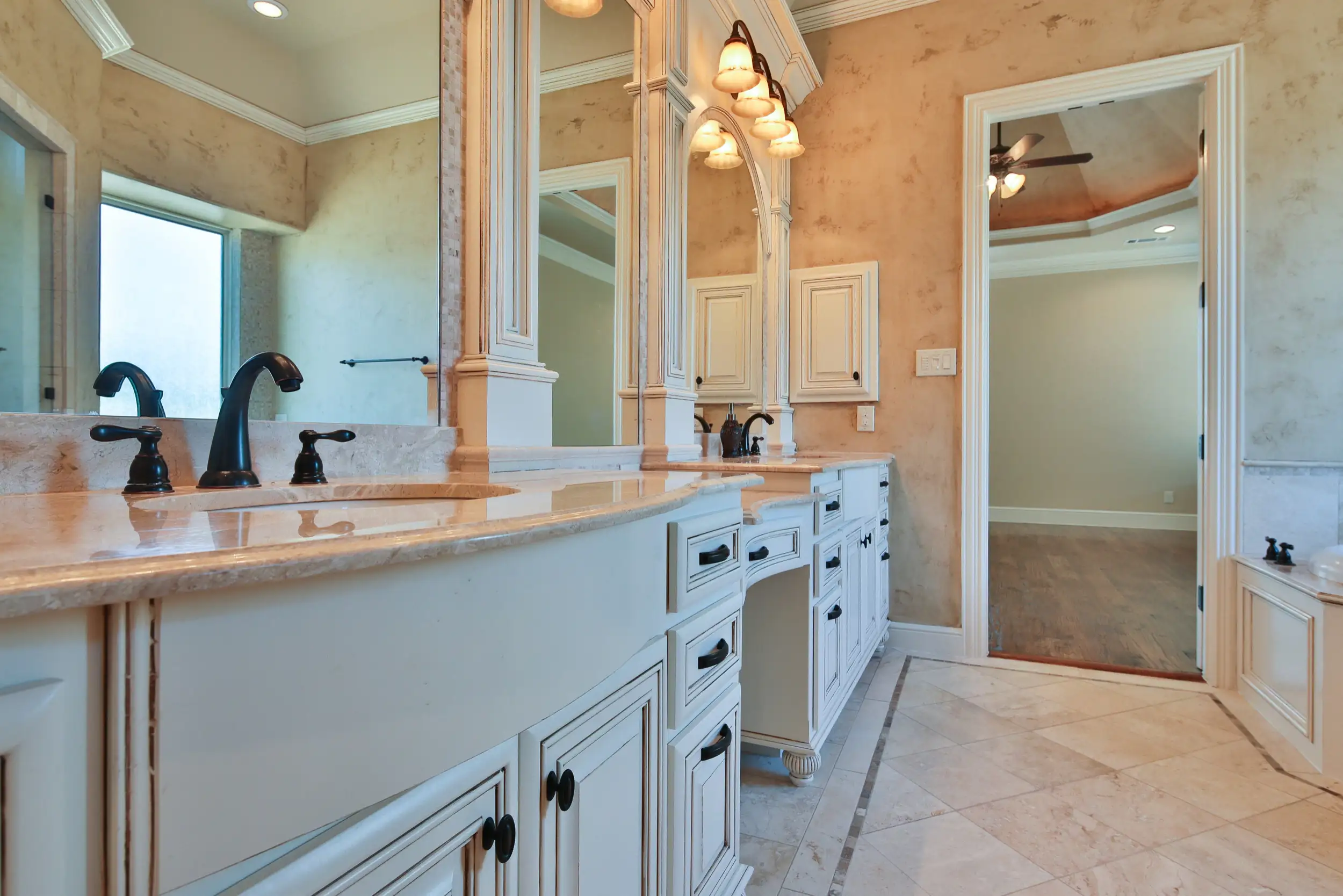 Elegant bathroom vanity with dual sinks, marble countertops, black fixtures, and ornate cabinetry, reflecting warm lighting in a large mirror.