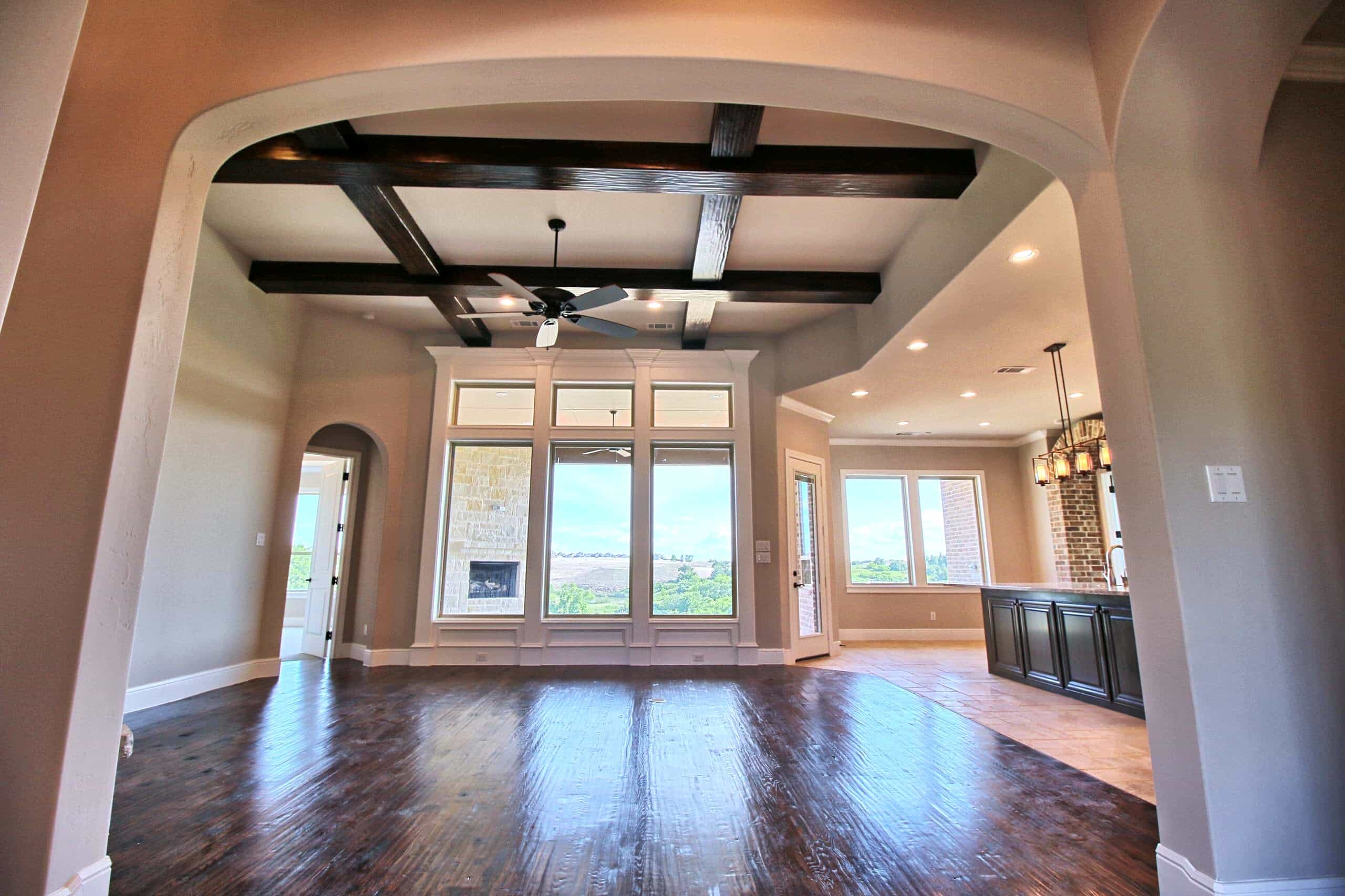 Living room seen through a wide arch, with dark wood ceiling beams, ceiling fan, hand-scraped hardwood floors and transom-topped windows facing a covered patio with stone fireplace, beside an open kitchen with dark island